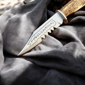 May include: A handcrafted knife with a polished metal blade and a serrated edge. The blade features etched text and is attached to a handle made of a light brown material. The knife rests on a gray fabric.