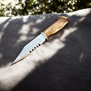 May include: A handcrafted knife with a polished silver blade and a serrated edge. The handle is made of natural antler, and the blade features etched text. The knife is set against a dark gray background.