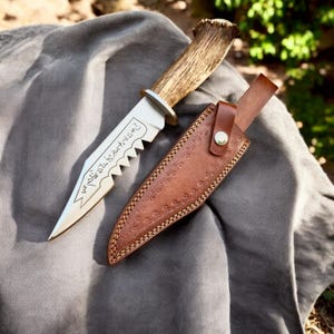 May include: A hunting knife with a serrated blade and a deer antler handle, resting on a gray cloth. The knife is accompanied by a brown leather sheath with decorative stitching.