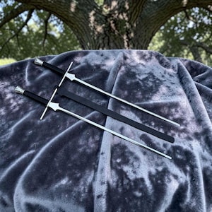 May include: Two silver-coloured metal swords with black handles and silver pommels, accompanied by a long, narrow black sheath. The swords feature straight, double-edged blades and cross-guards, positioned diagonally across the frame.