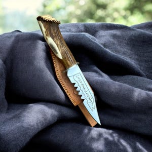 May include: A handcrafted knife with a blade etched with text and a serrated edge. The handle is made of antler, and the knife rests in a brown leather sheath. The knife is set against a dark gray fabric background.