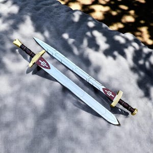 May include: Two toy swords with silver blades and brown handles are displayed on a gray surface. The hilts are gold with a burgundy emblem. One sword is in focus, while the other is slightly out of focus, both angled towards the top right.
