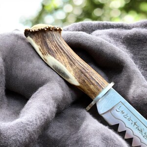 May include: Close-up of a knife with a handle made of antler and a silver blade. The blade has a serrated edge and etched text. The handle is brown and cream-colored, and the blade is silver.