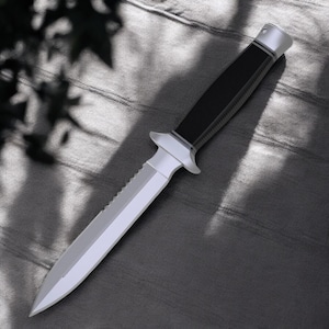 May include: A silver and black dagger. The blade has a serrated edge on one side. The handle is black with a textured grip. The dagger features a silver guard and pommel.