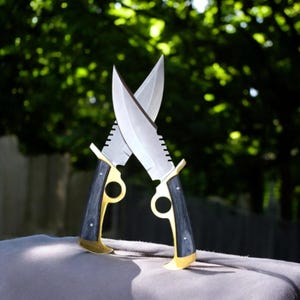 Può includere: Due coltelli incrociati con lame argentate e manici in legno nero. I manici sono impreziositi da metallo color oro e presentano un design circolare. Le lame sono seghettate vicino all'impugnatura.