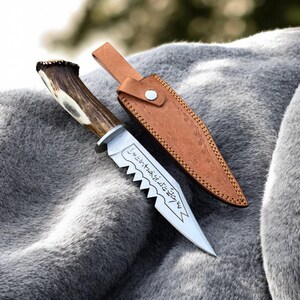 May include: A hunting knife with a serrated blade and a handle made of antler. The knife is accompanied by a brown leather sheath. The blade has etched text. The knife is resting on a gray, furry surface.