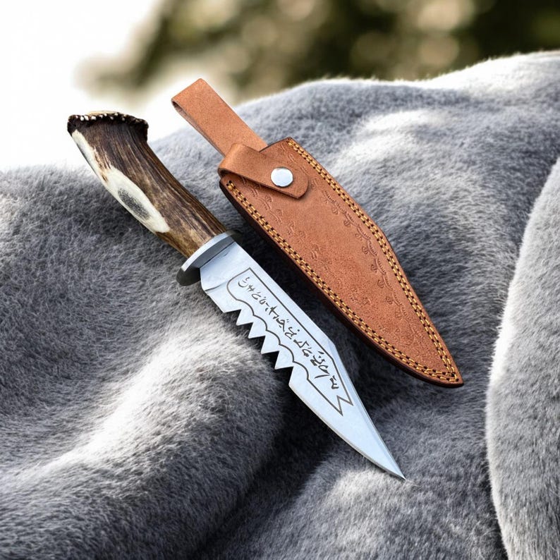 May include: A hunting knife with a serrated blade and a handle made of antler. The knife is accompanied by a brown leather sheath. The blade has etched text. The knife is resting on a gray, furry surface.
