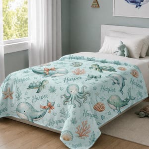 May include: A light blue blanket featuring a repeating pattern of watercolor-style sea creatures including whales, turtles, dolphins, and octopuses, interspersed with seashells, coral, and the name Harper printed in a cursive script font.