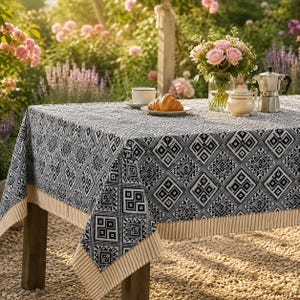 May include: A rectangular tablecloth featuring a black and white geometric pattern with a striped tan border. On the table sits a white teacup, a plate with a croissant, a glass vase with pink roses, a white sugar bowl, and a silver moka pot.
