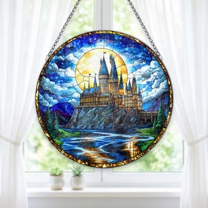 May include: Round stained glass window featuring a castle scene with a large yellow moon, blue sky, and water. The castle has multiple towers and is set on a rocky cliff. The window is framed with a dark border and a silver chain for hanging.