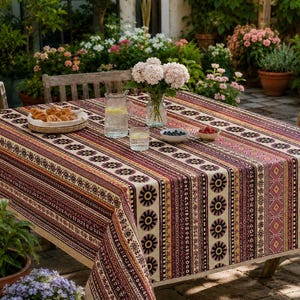 May include: A rectangular tablecloth featuring a repeating vertical pattern of dark red, orange, and cream stripes with stylised floral motifs. The fabric covers a wooden table set with a glass pitcher, two glasses, a bowl of croissants, and two small bowls.