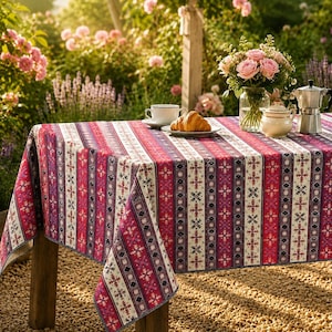 May include: A rectangular tablecloth featuring a vertical striped pattern in shades of red, pink, white, and dark blue, decorated with repeating geometric snowflake motifs. The fabric drapes over a table set with a croissant, cup, and coffee pot.