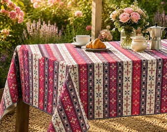 May include: A rectangular tablecloth featuring a vertical striped pattern in shades of red, pink, white, and dark blue, decorated with repeating geometric snowflake motifs. The fabric drapes over a table set with a croissant, cup, and coffee pot.