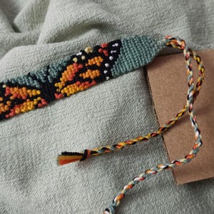 May include: A woven friendship bracelet featuring a detailed butterfly wing pattern in vibrant orange, black, and white threads, set against a sage green background. The bracelet includes long, braided multi-colored ties at one end for fastening.