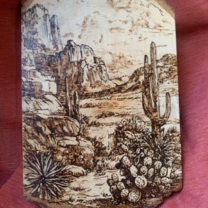 May include: A wood-burned art piece depicting a desert landscape with mountains, cacti, and other desert plants. The artwork is on a piece of wood with a natural edge, showcasing a detailed, brown-toned scene.