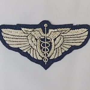 May include: A navy blue embroidered patch with a silver caduceus symbol and wings. The wings and the central staff are detailed with silver thread. The patch has a dark blue border. The medical emblem is centered.