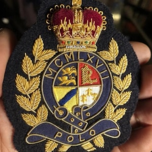Op de afbeelding: Een marineblauwe geborduurde patch met een gouden kroon, laurierbladeren en een schild met een gevierdeeld ontwerp. Het woord "POLO" staat op een blauwe banner. De patch heeft de tekst "MCMLXVI".