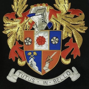 Op de afbeelding: Een geborduurd familiewapen met een zilveren helm, goudkleurige en rode dekkleden, en een schild verdeeld in vier secties met bloem-, gereedschap- en armmotieven. Onderaan staat een witte banner met TUTUS IN BELLO en de naam Rosenbaum eronder.