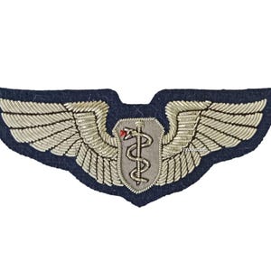 May include: A navy blue embroidered patch featuring silver wings and a central emblem with a medical symbol. The wings are detailed with silver thread, and the emblem includes a staff with a snake and a small red accent.