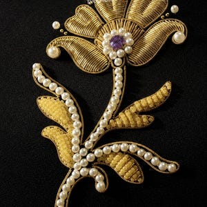 May include: A decorative floral patch featuring gold wire embroidery, white pearl beads, and yellow seed beads. The center of the flower has a purple crystal surrounded by small white pearls. The text EVO CHIC SMC is visible on the gold petals.
