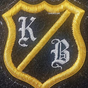 May include: Embroidered shield-shaped patch with gold trim and a diagonal gold stripe. The shield is divided into two sections, each with a silver letter: "K" and "B". The background is black. The text "EVOCHICMC" is visible.