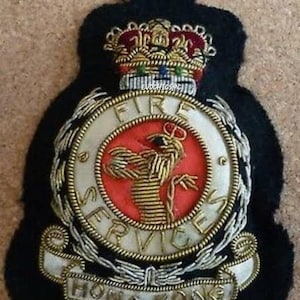 Może przedstawiać: Szczegółowa, haftowana naszywka z napisem "FIRE SERVICES HONG KONG". Naszywka ma czerwono-biały okrągły emblemat ze złotym wzorem feniksa. Korona znajduje się nad emblematem, a naszywka jest umieszczona na czarnym tle.