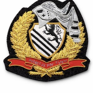May include: Embroidered patch with a heraldic design. The patch features a shield with black and white stripes and a black lion, surrounded by a gold wreath and a red banner with text. The patch is on a black background.