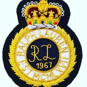 Puede incluir: Un parche bordado circular sobre un fondo azul marino oscuro con una corona dorada en la parte superior. El centro muestra el texto RALPH LAUREN EST MCMLXVII en hilo dorado alrededor de un círculo azul oscuro con las letras RL y 1967.