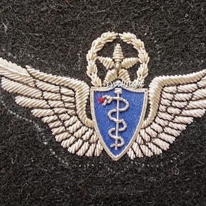 Op de afbeelding: Geborduurd embleem met zilverkleurige vleugels en een blauw schild met een medisch symbool. Boven het schild staan een ster en een krans. Dit insigne is waarschijnlijk voor militair of medisch personeel.