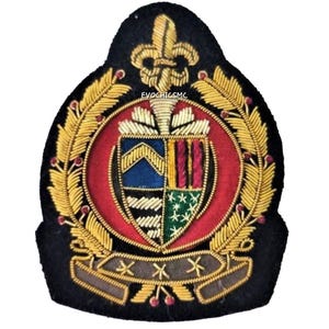 May include: A navy blue embroidered badge with a central shield design and a red border. The shield is divided into blue, white, green, and red sections. Gold-coloured laurel leaves and a fleur-de-lis decorate the top.
