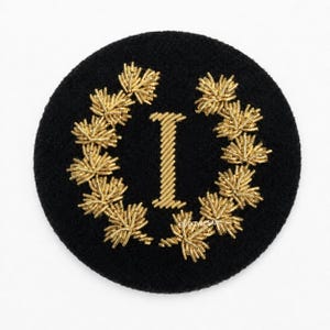 May include: A circular black patch featuring a central gold embroidered letter I surrounded by a wreath of gold metallic leaves. The design is crafted with intricate gold thread work, creating a textured appearance against the dark fabric background.