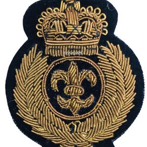 May include: A navy blue embroidered patch featuring a gold crown and a fleur-de-lis emblem. The patch is surrounded by gold laurel leaves and intricate gold thread detailing. The patch has a unique shape and design.