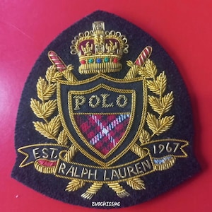 May include: A navy blue embroidered patch featuring a shield design with the word "POLO" at the top. The shield has a red and white plaid pattern. The patch is adorned with a gold crown, laurel leaves, and the text "EST. RALPH LAUREN 1967".