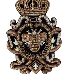 Pode incluir: Um emblema bordado preto com um motivo central de abelha rodeado por fio metálico dourado e bronze, strass transparentes e uma coroa no topo. O texto EVOCHICSMC está bordado em fio branco abaixo da coroa na base de tecido preto.