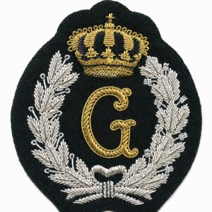 Op de afbeelding: Een zwarte geborduurde patch met een gouden kroon aan de bovenkant, een grote gouden letter G in het midden en een zilveren lauwerkrans rondom de letter. De tekst EVOCHICSMC is in kleine gouden letters onder de kroon geborduurd.