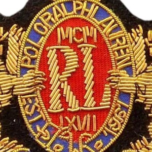 May include: A gold, red, and blue embroidered patch featuring a crown at the top and the text POLO RALPH LAUREN, MCM, RI, LXVII, and 17 REGENT STREET. The design includes intricate gold thread detailing on a black fabric base with a central oval.