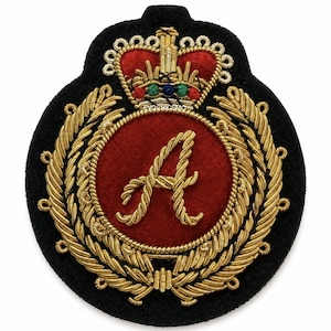 Puede incluir: Un parche bordado circular que presenta un círculo central de fieltro rojo con una letra A hecha de hilo de canutillo dorado. El diseño está rodeado por una corona de laurel dorada y rematado con una corona dorada detallada con acentos en rojo, verde y azul.