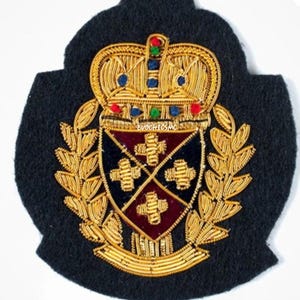 Op de afbeelding: Een zwarte stoffen embleem met gouden borduursel van een kroon boven een schild verdeeld in vier secties met gouden kruisen. Het schild is omringd door gouden laurierbladeren en de tekst EVO CHIC SMC is in het midden zichtbaar.