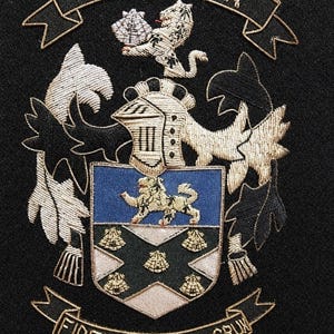 Könnte beinhalten: Ein gesticktes Wappen-Aufnäher auf schwarzem Hintergrund. Das Wappen zeigt einen Löwen, der eine Muschel hält, über einem Schild mit einem Löwen und fünf Muscheln. Ein Banner oben trägt die Aufschrift BAKER, unten steht FIDEI COTICULA CRUX.