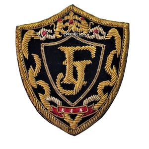 Op de afbeelding: Schildvormige geborduurde patch met een zwart midden en goudkleurige details. De patch heeft de letter "F" in goud, omgeven door sierlijke gouden borduursels en rode accenten. De tekst "EVOCHICSMG" is zichtbaar.
