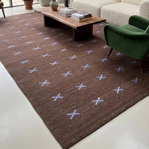 May include: A brown area rug with a repeating pattern of light blue crosses. The rug is rectangular and covers a large portion of a living room floor. The room features a white sofa, green armchair, and a wooden coffee table.