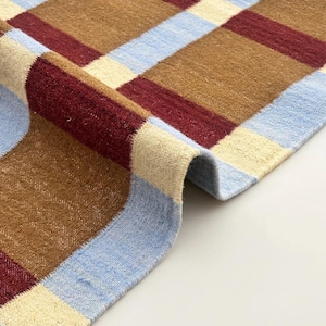 Könnte beinhalten: Nahaufnahme eines gewebten Teppichs mit Karomuster. Der Teppich hat Quadrate in den Farben Braun, Burgunderrot, Hellblau und Creme. Der Teppich ist unten aufgerollt und zeigt die Unterseite. Der Teppich besteht aus einem weichen, strukturierten Material.
