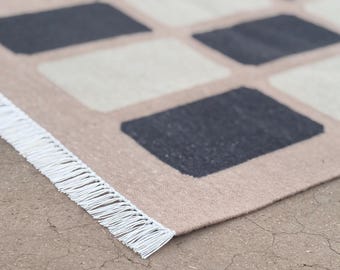 May include: Close-up of a beige rug with a geometric pattern of dark gray and off-white squares. The rug has a fringe of white threads along one edge. The rug is made of a woven material.