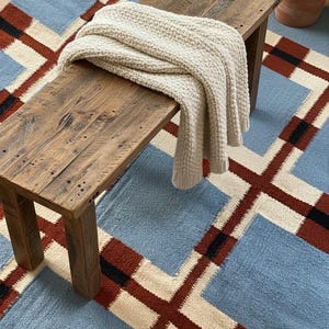 Op de afbeelding: Een rustieke houten bank met een crèmekleurige gebreide deken. De bank staat op een tapijt met een geometrisch patroon in blauw, rood, zwart en crème. Het patroon van het tapijt toont grote vierkanten en kruisen.