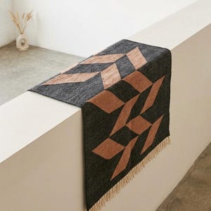 Op de afbeelding: Een rechthoekig tapijt met een geometrisch patroon van bruine en zwarte ruitvormen. Het tapijt heeft een franje langs de onderrand. Het tapijt wordt op een wit oppervlak gepresenteerd.