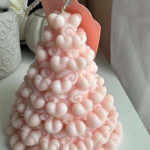 Puede incluir: Una vela rosa pálido con forma de árbol de Navidad, adornada con detalles en forma de corazón. La vela tiene una superficie texturizada y está sujeta a una etiqueta rosa con una cuerda.