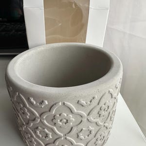 May include: A grey, round concrete planter with a detailed floral pattern. The planter has a wide opening and a textured surface. A white box is in the background.
