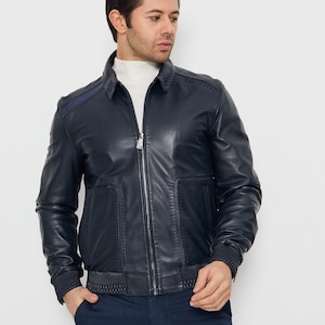 May include: A navy blue leather jacket with a zip-up front, a collar, and ribbed cuffs. The jacket has two front pockets and a quilted design on the shoulders. The model is wearing a white roll neck and navy blue trousers.