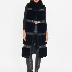 May include: A long, black faux fur vest with horizontal leather-like bands. The vest has a high collar and is worn over a black top and black pants. The model is also wearing black, knee-high, heeled boots. The background is white.
