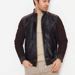 May include: A black and brown leather bomber jacket with a zip-up front. The jacket has brown sleeves and a textured shoulder panel. The model is wearing a beige turtleneck and tan trousers.